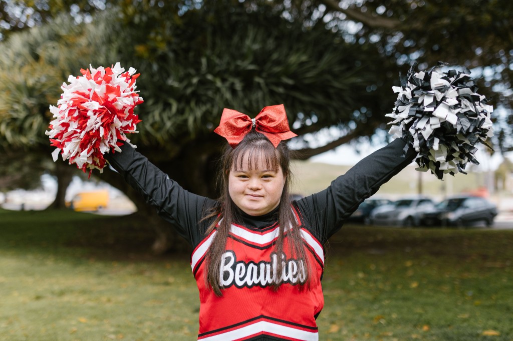 A smiling young woman with Down syndrome in a cheerleader uniform, raising pom-poms outdoors